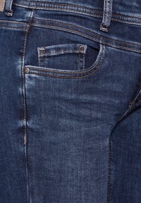 Jean en denim bleu foncé avec un design classique à cinq poches, détails de couture visibles et un petit rivet en métal accentué sur la poche arrière.