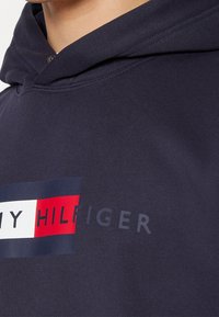 Gros plan d'un sweat à capuche bleu marine montrant le logo Tommy Hilfiger avec un design rectangulaire rouge, blanc et bleu sur la poitrine.