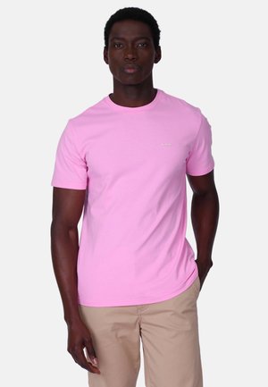 Homme portant un t-shirt à manches courtes rose clair uni et un pantalon beige, debout avec une main dans la poche, sur un fond blanc.
