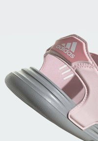 Calzature Adidas rosa con una toma superiore testurizzata, design con tacco aperto e una suola grigia flessibile caratterizzata da tre scanalature. Dettagli del logo bianco visibili.