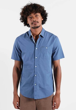 Homme aux cheveux bouclés portant une chemise à manches courtes bleue boutonnée et un pantalon marron, debout devant un fond blanc uni.