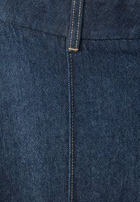 Gros plan sur un tissu en denim bleu foncé montrant une couture verticale et une boucle de ceinture avec des surpiqûres jaunes.