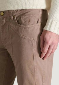 Pantalones de algodón marrón claro con una textura suave, que presentan un detalle destacado en el bolsillo y un parche con logo. Incluye herrajes de botón dorado.