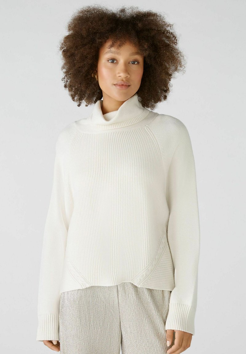 Weißer, gerippter Strickpullover mit hohem, umgeschlagenem Kragen und langen Ärmeln, der eine lockere Passform und dezente Seitendetails aufweist.