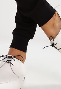 Chaussures blanches en cuir avec un bout arrondi, lacets noirs et profil bas, accompagnées de pantalons noirs ajustés. Texture lisse, design minimal.