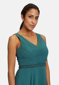 Ujjatlan türkiz ruha plisszírozott felsőrésszel, V nyakkivágással és gyöngyökkel díszített övvel. Simább anyagtextúra. Elegáns sziluett.