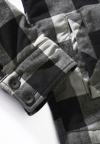 Juodas ir pilkas languotas audinys su švelnia tekstūra, kuriame matomas artimas marškinių rankogalių vaizdas, du juodi mygtukai ir vienos kišenės detalė.