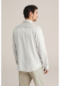 Mann mit lockigem dunklem Haar, der ein weiß gestreiftes Langarmhemd und beige Hose trägt, seitlich vor einem einfarbigen Hintergrund stehend.