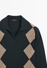 Donkergroen gebreide trui met een V-hals en een patroon van grote beige diamanten, met een zachte textuur en een label bij de kraag.