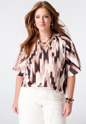 Femme aux longs cheveux roux portant un chemisier à manches courtes à motifs, un pantalon blanc, des colliers superposés, des boucles d'oreilles créoles et plusieurs bracelets.