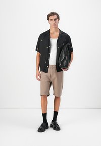 Ung mann iført svart blomstrete kortermet skjorte, hvit tank topp, beige shorts, svarte sokker og sko, holder en stor svart skinnveske.