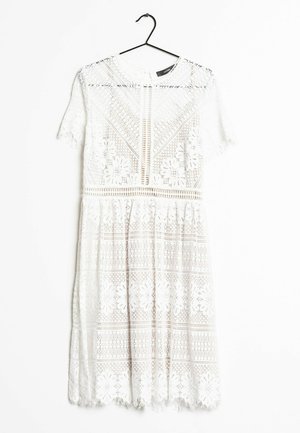 Robe blanche en dentelle jusqu'aux genoux avec motifs floraux, manches courtes, encolure ronde, et détails verticaux à l'avant, suspendue à un cintre noir.