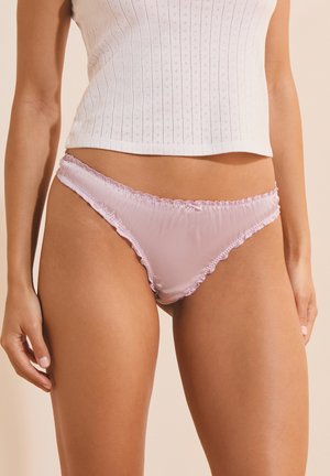 Femme portant un ensemble de lingerie bikini en satin rose clair et un crop top blanc sans manches en maille avec un petit motif œillet.