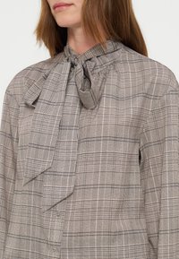 Femme portant un chemisier beige à carreaux avec un grand nœud noué au col et des manches longues.