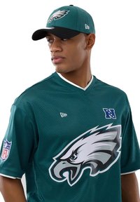 Homme portant un maillot vert des Philadelphia Eagles et une casquette assortie, regardant sur le côté avec un fond blanc.