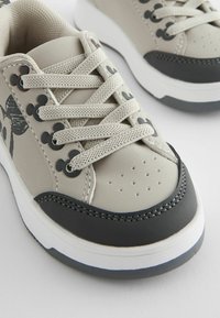 Beige sneakers with black accents, featuring a textured upper, perforated toe, and contrasting laces. Notable graphic design detail on the side.