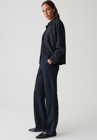 Zwarte button-up shirt over een donkere top, gecombineerd met marineblauwe wijde broek. Gladde textuur, aangesloten stijl en zwart instap schoenen.