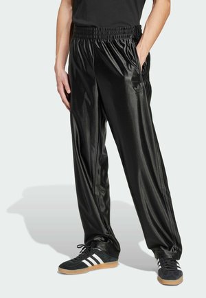 Personne portant un pantalon de survêtement noir brillant avec une taille élastique et des baskets noires avec des bandes blanches, debout avec une main dans la poche.