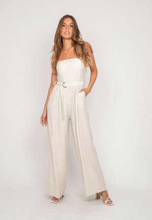 Femme debout, une main dans la poche, portant une combinaison blanche sans bretelles avec ceinture et des sandales à talons blanches sur un fond clair.