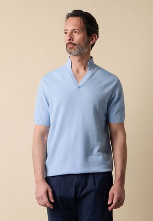 Mann mit kurzen dunklen Haaren und Bart, trägt einen hellblauen kurzärmligen Pullover mit V-Ausschnitt und dunkle Hose, steht vor beigem Hintergrund.