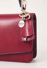 Borsa a tracolla in pelle color berry con texture liscia, hardware dorato e un'etichetta con portachiavi. Presenta linee pulite e cuciture a contrasto.