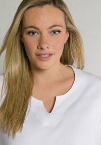 Top bianco con scollatura a V e una superficie liscia. Presenta un delicato dettaglio di cucitura; indossato da una persona con capelli lunghi e lisci biondi.