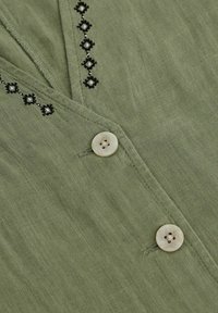 Blusa verde oliva realizzata in tessuto leggero con dettagli ricamati neri e chiusura con un solo bottone crema. Texture liscia.