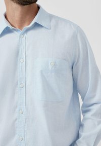 Jasnoniebieska, długorękawowa koszula zapinana na guziki, wykonana z teksturowanej tkaniny. Posiada kieszeń na piersi z guzikiem oraz standardowy kołnierz.