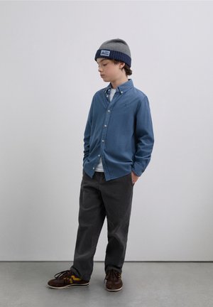 Camisa azul claro abotonada sobre una camiseta blanca, gorro gris, pantalones de pana grises y zapatos marrones con detalles en amarillo.