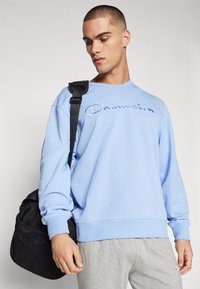 Hellblaues Sweatshirt mit gesticktem "Champion"-Logo, langen Ärmeln, gerippten Bündchen, kombiniert mit grauen Jogginghosen und einer schwarzen Tasche.