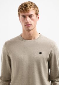 Beige gebreide sweater met een ronde halslijn, textuurstof en een klein zwart hexagonaal logo op de linkerborst. Lange mouwen, aansluitend ontwerp.