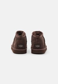 Bocanci scurți din suede maro, cu căptușeală pufoasă, talpă din cauciuc plat, curele de prindere pe spate și un mic logo UGG pe fiecare toc.