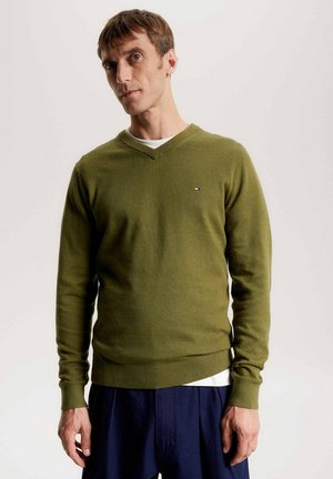 Suéter de algodón verde con cuello en V y mangas largas. Presenta un pequeño logo de la marca en el lado izquierdo del pecho. Combinado con pantalones azul oscuro.