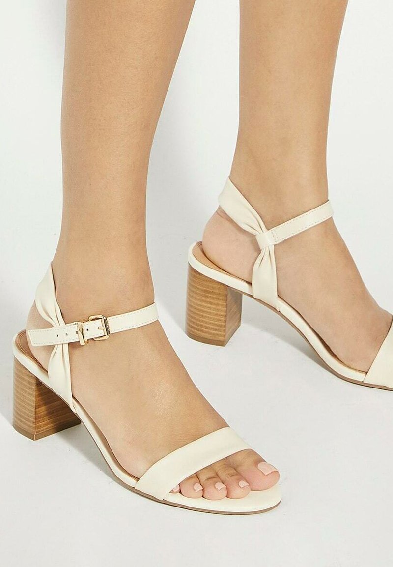 Talons ouverts couleur crème avec un bloc en bois, une bride ajustable à la cheville, et un détail en cuir noué au talon. Texture lisse.