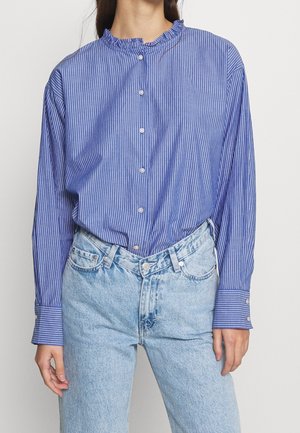 Blau-weiße vertikal gestreifte Button-up-Bluse mit einem Rüschkragen und langen Ärmeln, kombiniert mit hellblauen Jeans.