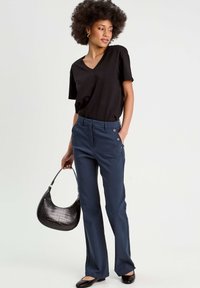 Schwarzes V-Ausschnitt-T-Shirt, maßgeschneiderte navyblaue Hose mit seitlichen Knöpfen und schwarze Handtasche mit Kroko-Prägung. Das Model trägt schwarze Ballerinas und hat eine aufrechte Haltung.