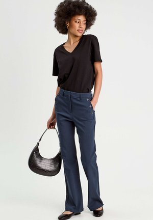 T-shirt noir en v, pantalon tailleur bleu marine avec boutons sur les côtés, et sac à main noir gaufré crocodile. Le mannequin porte des ballerines noires et adopte une posture droite.