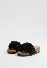 Chanclas negras con suela de corcho en forma de cuña, que presentan grandes adornos florales negros en las correas y una textura mate suave.