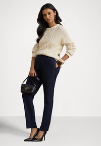 Camisola creme de malha com cabo, punhos canelados, calças de alfaiate azul-marinho, saltos altos pretos e uma pequena mala preta com ferragens douradas.