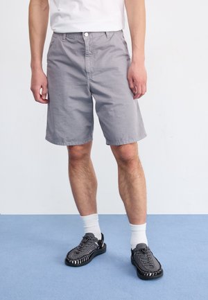 Mann trägt hellgraue kniehohe Shorts, weiße Socken und schwarze gewebte Schuhe, steht auf blauem Boden vor weißem Hintergrund.