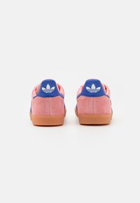 Pink suede sneakers with blue accents, featuring a textured gum sole and white logo on the heel. Rounded shape with a low profile design.