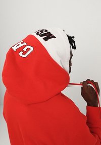 Persona con sudadera roja y blanca con el texto "GAP" y "MSG" en la capucha, tirando del cordón, de perfil frente a un fondo liso.