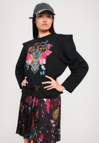 Sudadera negra con bordado de flores y mariposas en colores, con hombros estructurados. Combinada con una falda floral plisada y cintura ceñida.