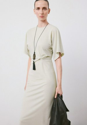 Femeie purtând o rochie bej mulată cu detaliu legat și un colier cu ciucuri negri, ținând o jachetă de piele neagră pe un fundal alb.