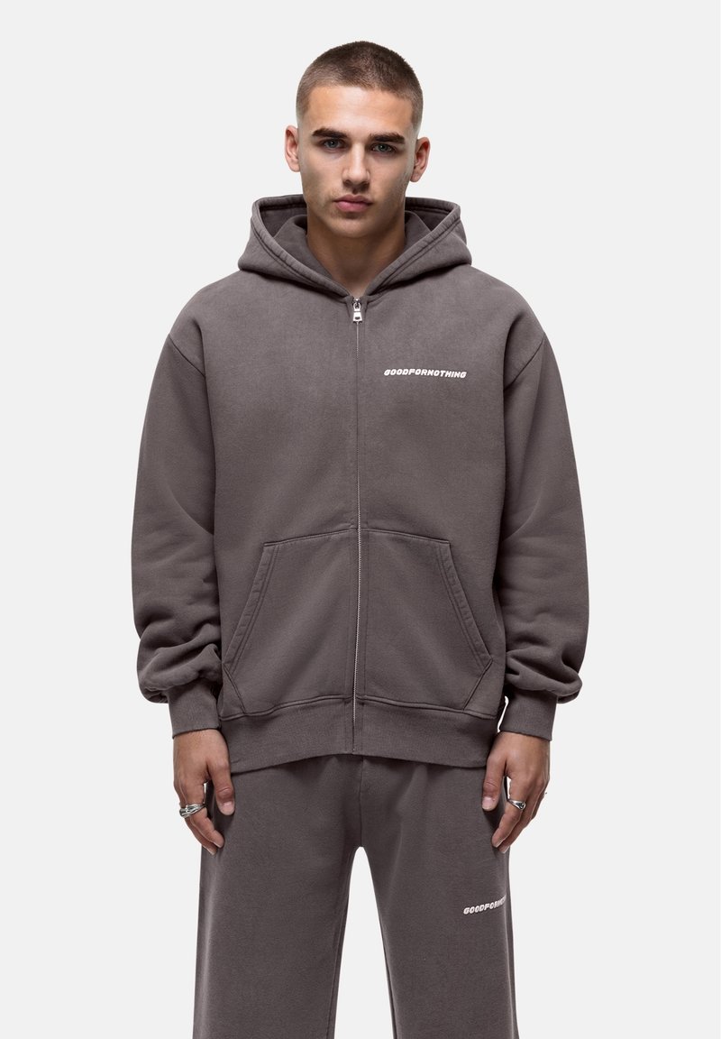 Grijze zip-up hoodie met een voorkantzak, gemaakt van zachte stof. Heeft een logo op de linkerkant van de borst. Draagt samen met bijpassende grijze broek.