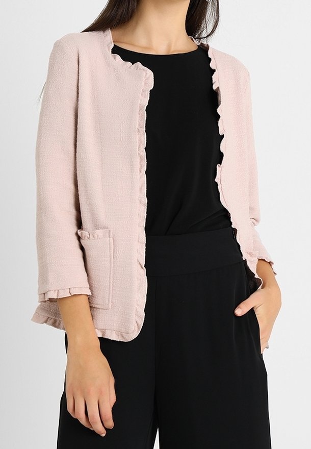 Femme portant une veste rose clair texturée à ouverture sur le devant avec des bords à volants, par-dessus un haut noir et un pantalon noir, une main dans la poche.