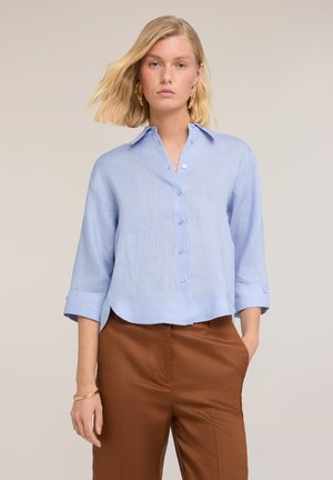 Blonde Frau mit goldenen Ohrringen, hellblauer Bluse mit Knöpfen und hochgekrempelten Ärmeln sowie brauner Hose mit hohem Bund, eine Hand in der Tasche.