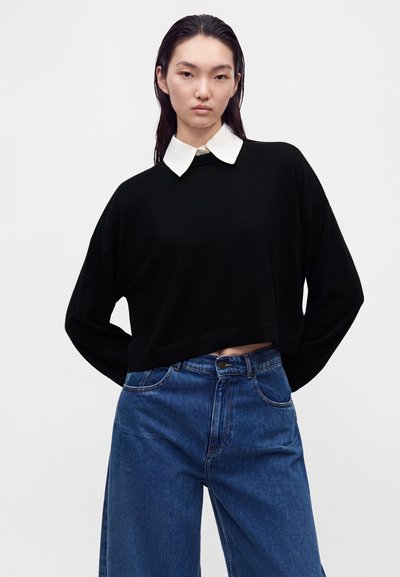 Jeune personne aux longs cheveux foncés portant un pull noir avec un col blanc et un jean bleu taille haute, debout les mains dans le dos.