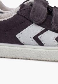 Dark suede sneaker with white accents, featuring a chevron design, and two Velcro straps. The textured sole provides added grip.