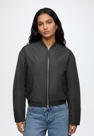 Mujer con cabello largo y oscuro, vistiendo una chaqueta bomber negra con cremallera y vaqueros azules, de pie frente a un fondo blanco liso.